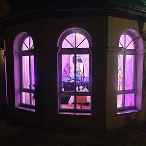 Blick durch Fenster auf festlich beleuchtete Hochzeit mit DJ Greußen. Die ART of SOUND als DJ Greußen sorgt mit stilvoller Beleuchtung und professionellem DJ-Pult im Innenraum für romantische Atmosphäre bei einer Hochzeitsfeier. Außenaufnahme bei Nacht mit violettem Lichtschein.